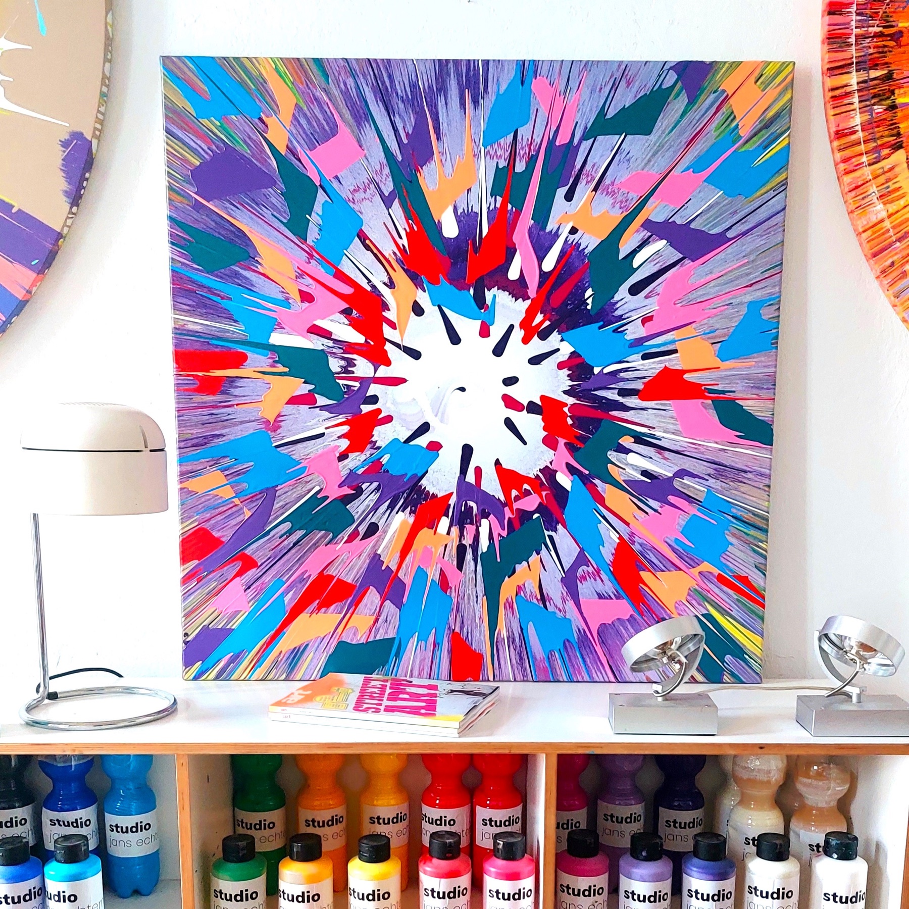 Quadratisches Spin Painting in Blau, Violett, Pink und Rot über einem Farbregal im Studio