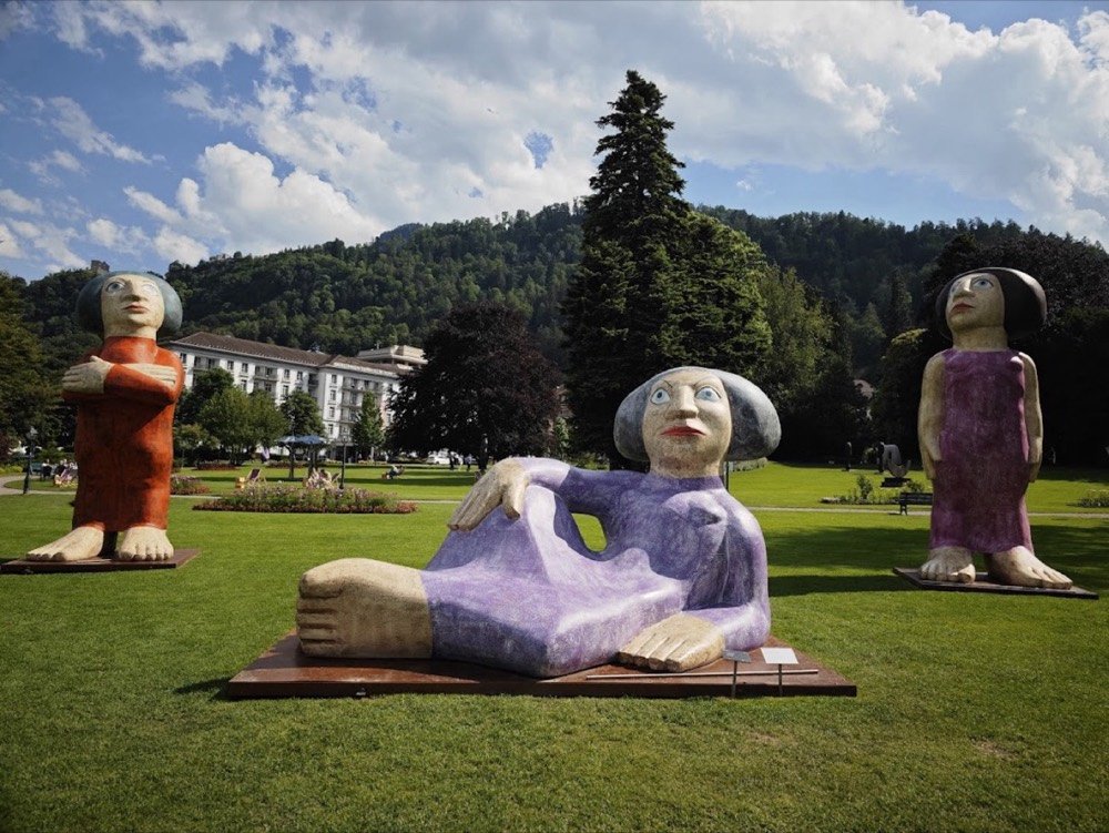 Große Figurenskulpturen der Bad Ragartz auf einer Wiese, im Hintergrund Hotel und Berglandschaft