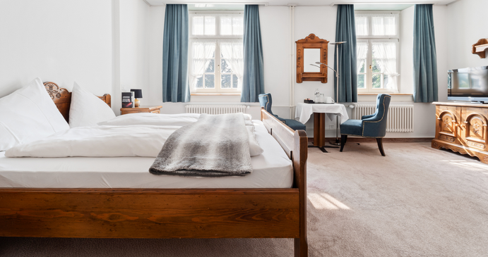 Helles Hotelzimmer mit Holzbett, Sitzecke mit zwei blauen Sesseln am Fenster und Holzmöbeln